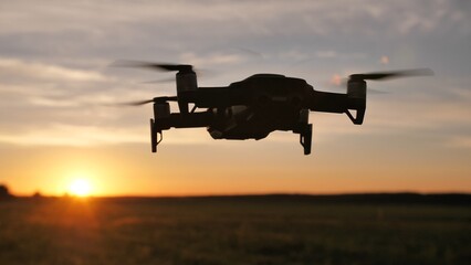 Drone flies gracefully over a vast field at sunset, its propellers spinning in the orange sky, creating a modern silhouette against the rural landscape