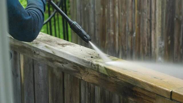 A man cleans a deck railing with a pressure washer sprayer