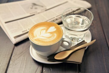 A latte and a glass of water on a table with a newspaper, suggesting a relaxing moment.