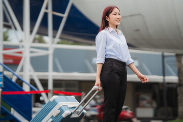 Businesswoman Arriving at Airport: A young, stylish businesswoman with a confident smile walks towards the airport terminal, pulling her luggage, ready to embark on a journey. The photo captures a sen © Nittaya