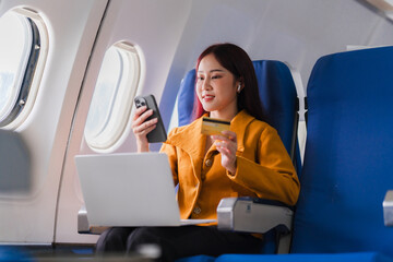 In-Flight Shopper: A young Asian woman enjoys the convenience of online shopping, browsing on her phone and holding a credit card while seated comfortably in an airplane cabin. 