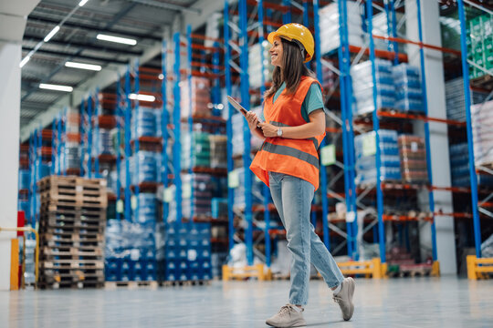 Woman warehouse supervisor with helmet and clipboard walking, inspecting.