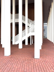 Naklejka premium Large white columns supporting a modern style building. There's an exterior stairway to the second floor. The uneven ground is covered with red brick. The stairway is metal and painted white. 
