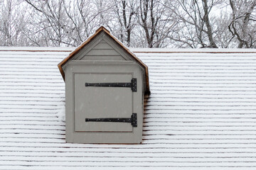 A tan colored vintage building with a pitched roof, a steep slope, cedar shakes, window dormer and a wood shutter. There are black metal hinges on the shutter.The roof is covered with fresh white snow