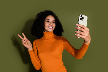 Photo of cheerful pretty lady dressed orange turtleneck showing v-sign tacking selfie gadget isolated green color background