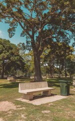 Obraz premium A photo of a concrete bench in a park. The bench is situated near a tree, with lush green leaves. There's a path leading to the bench. The background contains other benches and trees. 