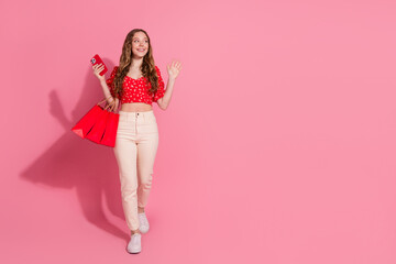 Full body photo of teen woman look empty space walk wave hand shopping device dressed stylish red clothes isolated on pink color background