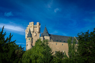 Fototapeta premium View of the north face of the Alcazar of Segovia, Castilla y Leon, Spain, from the Paseo del Eresma with its thick grove of trees and intense blue sky
