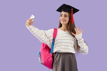 Happy female student in graduation hat with backpack taking selfie on purple background