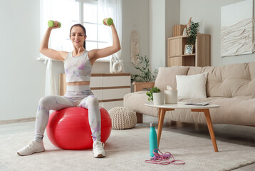 Fototapeta premium Sporty young woman with dumbbells training on fitball at home