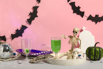 Glass of cocktail, tableware and decorations for Halloween on table against pink background