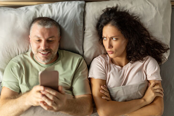 Couple in bed. Unsatisfied angry wife frustrated while her internet addict husband using mobile phone ignoring her, above view