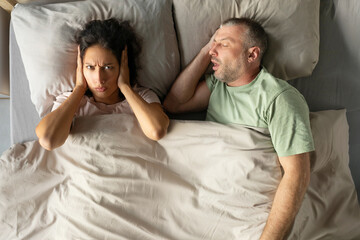 Annoyed woman closing ears with hands suffering from loud snore of her sleeping husband lying...
