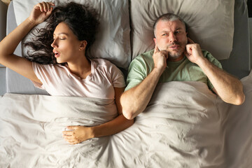 Disturbed man covering her ears with fingers to block out the loud snoring of wife sleeping nearby,...