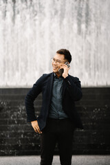 Businessman on the Go: A young, stylish businessman in a navy blazer and glasses smiles as he takes a phone call, showcasing a sense of professionalism and connection. The backdrop of a modern urban f