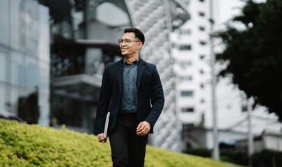 Urban Commute, Confident Future: Young Asian professional strides through city with laptop, ambition in his stride. 