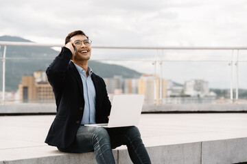 Urban Professional on the Go: A young Asian businessman enjoys a productive phone call on a rooftop, laptop in hand, with a vibrant cityscape as his backdrop. 