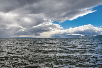 A sky full of beautiful clouds, lit by the sun. Before that it was raining. The rippling waters of the lake overflow in the light. Mountains in the distance.