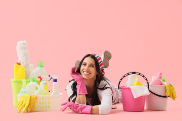 Beautiful housewife with cleaning supplies lying on pink background