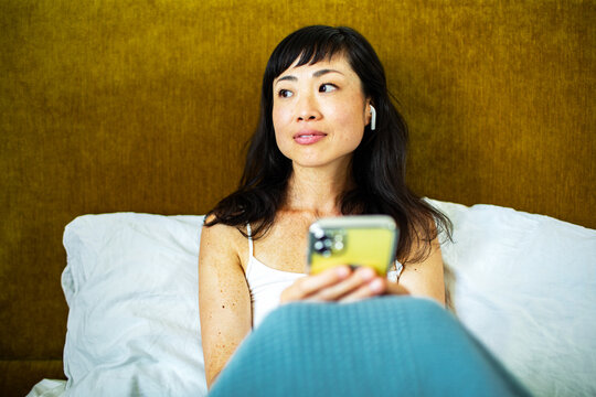 Smiling Asian woman using smartphone in bed at home