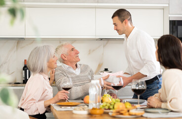 Adult son hands gift, box tied with ribbon, to elderly dad during feast, family holiday dinner. Senior spouses celebrate event in cozy family atmosphere in company of younger couple