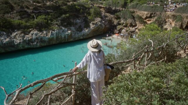 Woman overlooking stunning cala del moro bay in mallorca, spain on a sunny day, surrounded by nature, cliffs, and turquoise waters.