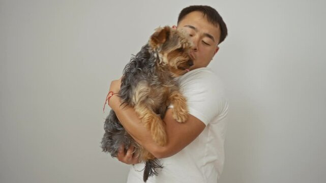 A young asian man from china holds his small pet dog affectionately against a white isolated background wall.