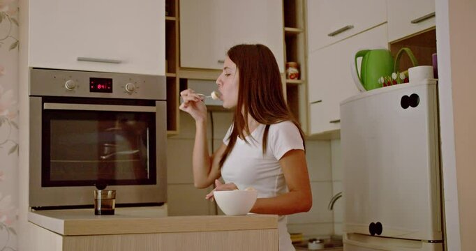 A young woman eats breakfast and drinks coffee in her kitchen. She is enjoying her morning and dancing while she waits.