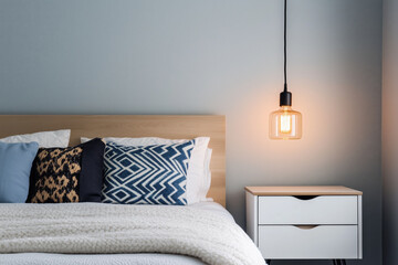 A modern bedroom featuring a cozy bed with patterned pillows, a warm pendant light, and a minimalist nightstand against a light-colored wall