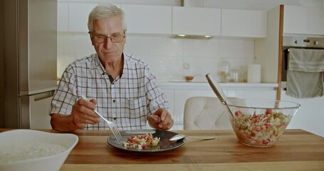 An elderly man sits at his kitchen table and enjoys a healthy lunch of salad. He's happy to eat his fresh food and talks to family on a video call.