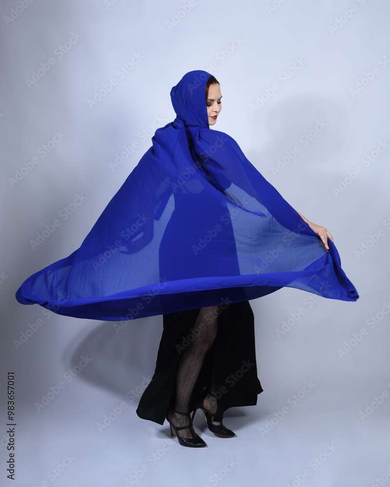 Sticker Full length portrait of  female red head model wearing  glamorous black witch dress with blue fantasy cloak halloween costume.  standing pose with gestural arms, isolated figure studio background.