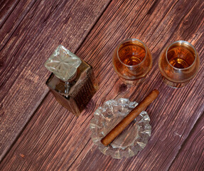 A crystal decanter and two glasses of expensive cognac and a glass ashtray with a cigar on a dark wooden table.