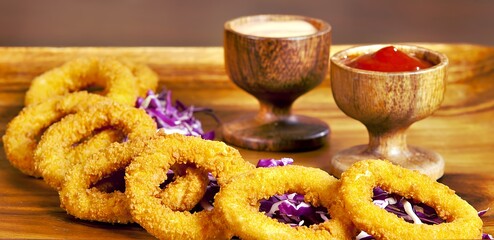 A wooden platter featuring crispy onion rings with two dipping sauces and purple cabbage.