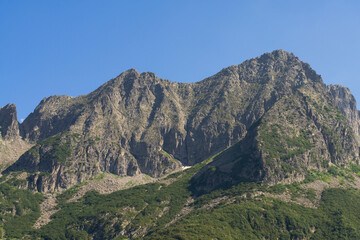 Striking view of rugged mountain peak, with rocky textures and sparse vegetation, set against a clear blue sky, highlighting the raw beauty of nature