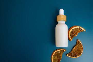 Flat lay of a white dropper bottle with wooden cap on a deep blue background with dried oranges