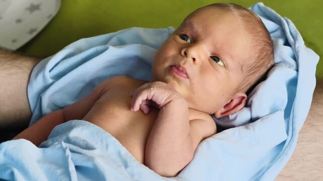 Close up of an infant. A newborn baby wrapped in a light blue nappy in the arms of a parent. The skin on the baby's hands is peeling. There are signs of mild jaundice