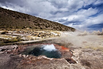 A geothermal hot spring bubbling amidst a rugged landscape under a cloudy sky.