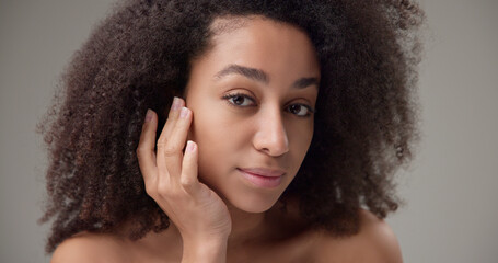 Beauty and healthcare concept - beautiful African American woman with curly afro hairstyle and clean, healthy skin touches her cheek and face with her hand, posing and looking at the camera