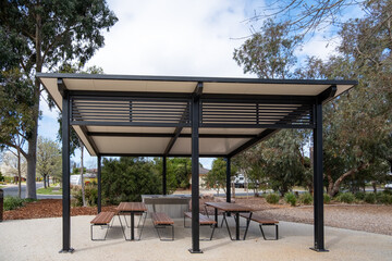 A covered picnic shelter in a public park, with tables and benches and a barbecue grill, in a suburban neighborhood in Australia. Parkland community amenities and recreational facilities.