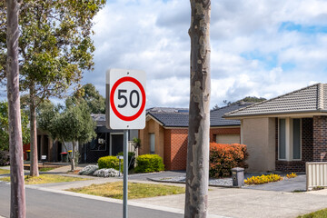 A speed limit sign indicating a 50 km per hour zone warns car drivers to slow down for residents' safety, located on a suburban street with a row of Australian residential houses in the neighborhood.