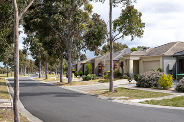 A quiet, clean, and well-maintained suburban street is lined with trees and modern, single-story houses in an Australian residential neighborhood in a suburb.