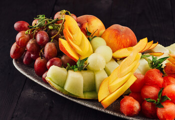 Oval plate overflowing with fresh watermelon, cantaloupe, peaches and grapes on dark wooden table