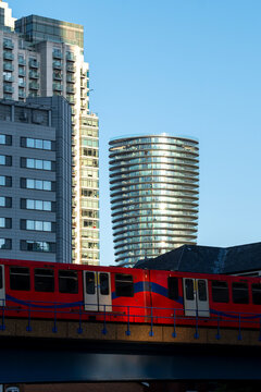 A bright red and blue train passes by towering skyscrapers in an urban setting during daylight, showcasing transportation, modernity, and city life.
