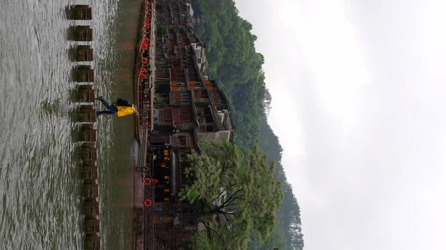 Tracking shot of a man in yellow raincoat crosses Tuojiang on Stepping Stones in Fenghuang on a cloudy day, China. Vertical