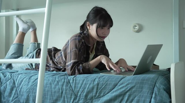 Asian girl prone on bed and typing on laptop