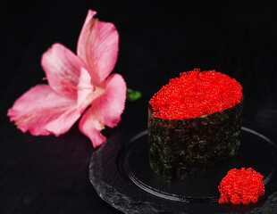 Gunkan maki sushi with red caviar tobiko and pink flower resting on black slate plate © AntAlexStudio