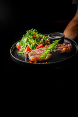 Grilled steak garnished with fresh herbs and tomatoes, served on a black plate with sauce, against a dark background. Perfect for culinary, food photography, and restaurant menu concepts.