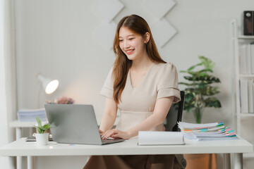 Focused Productivity: A young Asian woman beams at her laptop, engrossed in work at her bright, minimalist home office. 