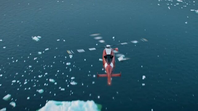 Footage of a orange rescue helicopter flying over some arctic ocean sea water filled with white ice chunks. Water is dark blue and has small ripple waves. Helicopter preforms a turn and flies straight