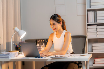 Focused and Determined: A young Asian woman, radiating determination, sits at her desk, her eyes focused on her laptop, late into the night, showcasing dedication and the pursuit of goals. 